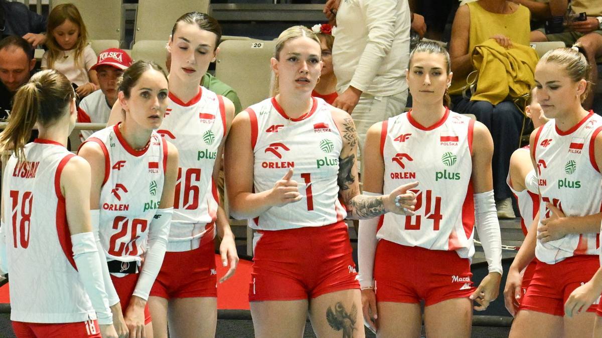 Grupa zawodniczek drużyny siatkarskiej w białych koszulkach z czerwonymi detalami i czerwonych spodenkach, stojących w szeregu na tle trybun.
