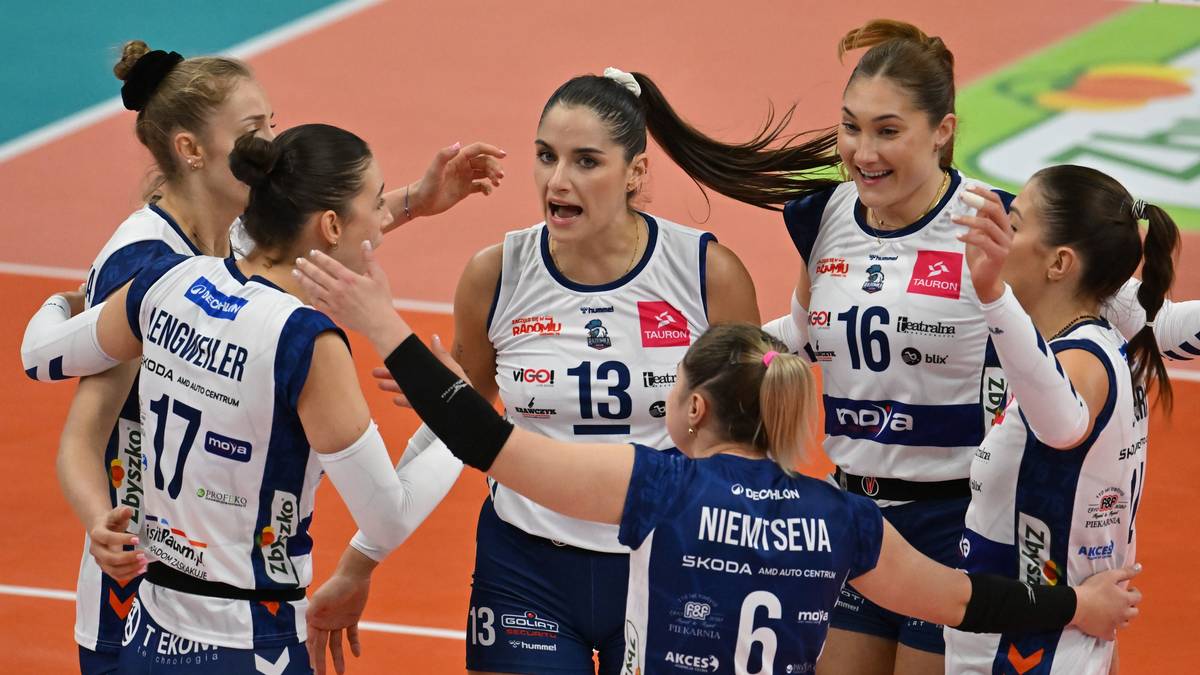 Grupa sześciu siatkarek w białych strojach z niebieskimi wstawkami, cieszących się ze zwycięstwa na boisku.