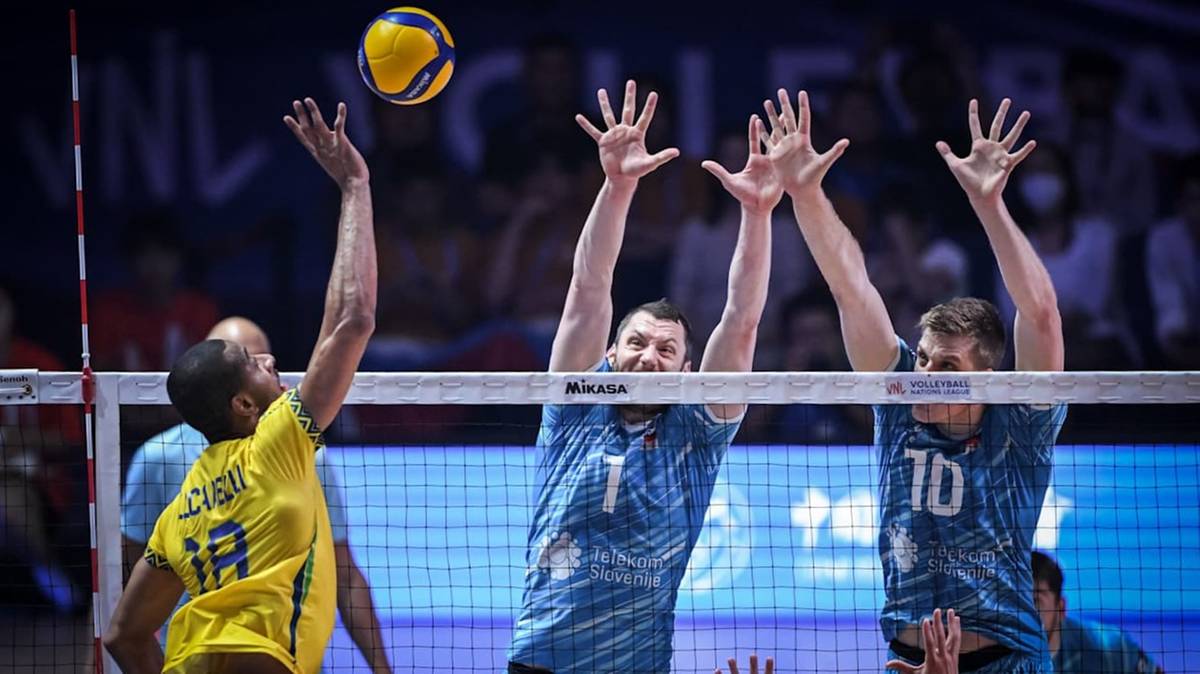 Siatkarz w żółtej koszulce wykonuje zagrywkę, podczas gdy dwaj gracze w niebieskich koszulkach próbują zablokować piłkę.