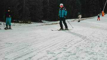 Osoba na nartach zjeżdżająca po ośnieżonym stoku narciarskim, w tle inne osoby na nartach i drzewa.