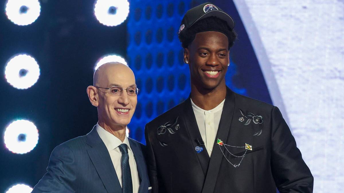 Adam Silver, komisarz NBA, w garniturze i z okularami, podaje rękę Darrynowi Petersonowi, młodemu koszykarzowi w eleganckim czarnym stroju i czapce z daszkiem, podczas ceremonii draftu NBA. Obaj się uśmiechają.