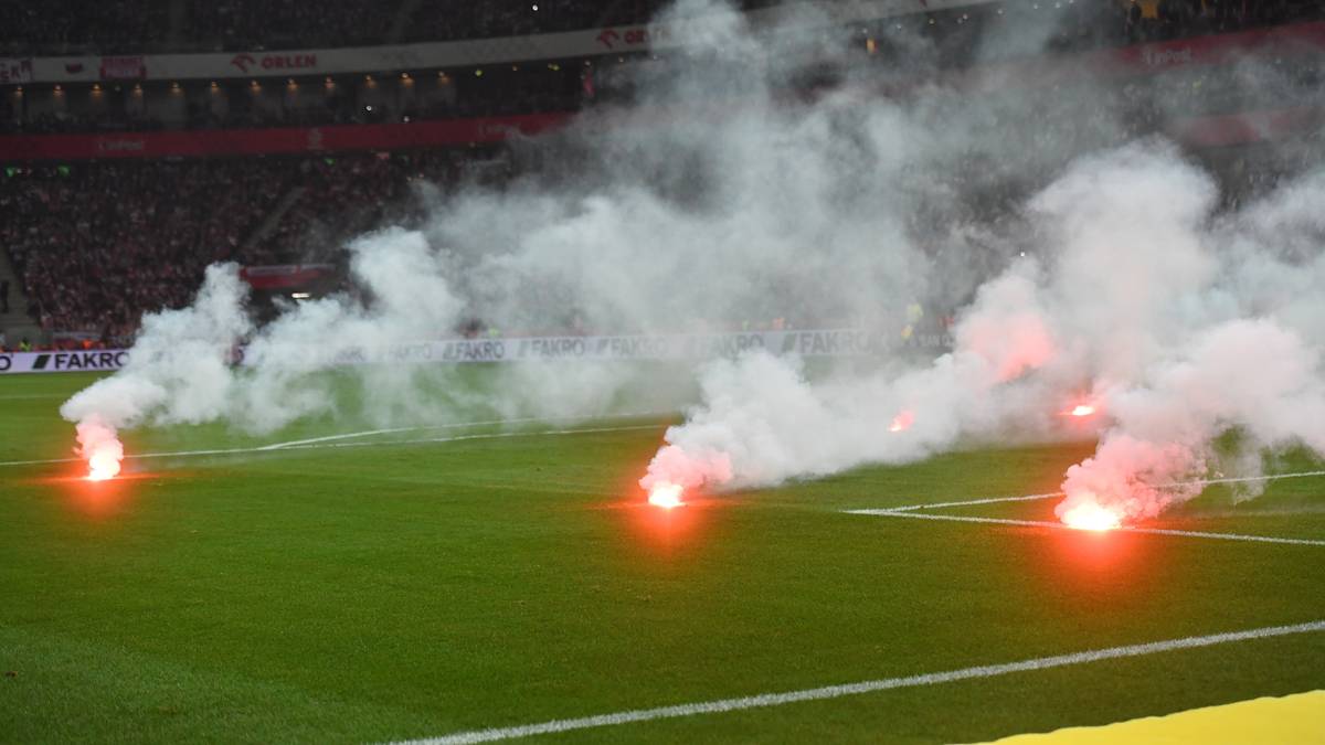 Na boisku piłkarskim płoną race, z których unosi się biały dym i czerwone światło.