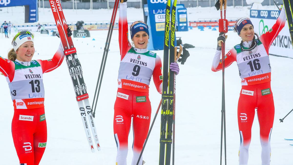 Trzy zawodniczki narciarstwa biegowego w czerwonych strojach sportowych stoją obok siebie, trzymając narty i kije. Dwie zawodniczki po bokach unoszą ręce w geście radości. Na ich strojach widoczne są numery startowe.