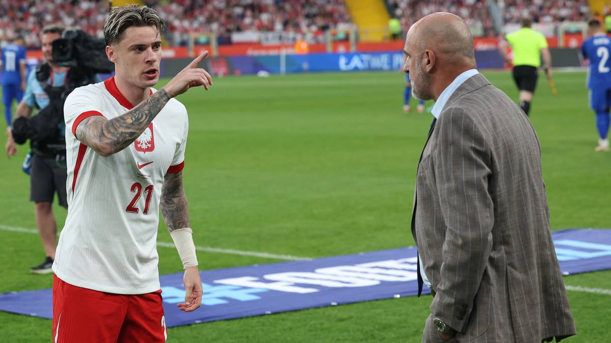 Piłkarz reprezentacji Polski w białej koszulce z numerem 21 rozmawia z starszym mężczyzną w garniturze na boisku piłkarskim.