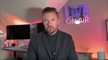 Mężczyzna w czarnej marynarce i golfie patrzy prosto w kamerę. Za nim znajduje się biurko z monitorem komputerowym i klawiaturą, a na ścianie neonowy napis "LIVE ON AIR" w kolorze fioletowym. Tło jest oświetlone czerwonym światłem.