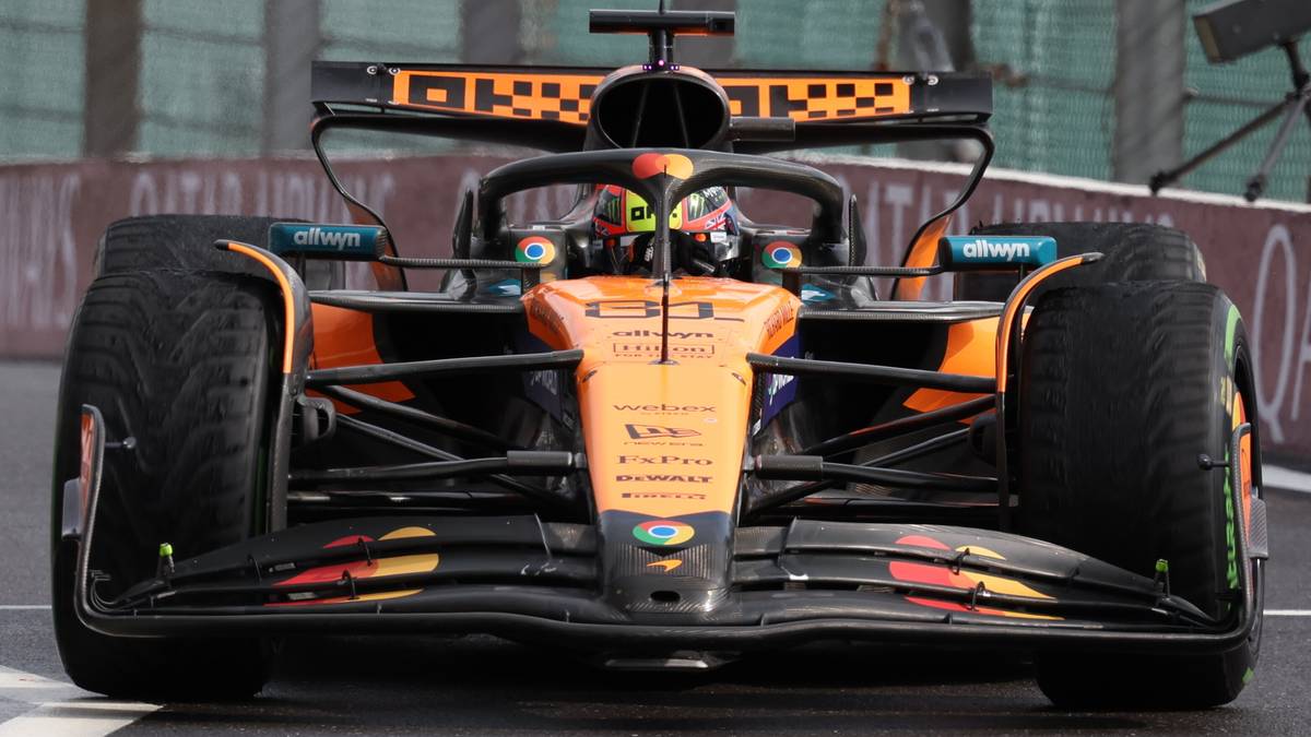 Widok z przodu na bolid Formuły 1 zespołu McLaren z kierowcą w kasku, w barwach pomarańczowo-czarnych, na torze wyścigowym.