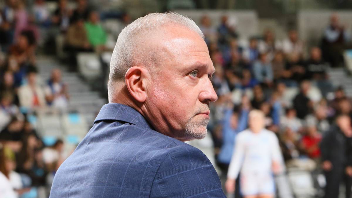 Portret mężczyzny w niebieskiej koszuli z krótkim rękawem, z siwymi włosami, patrzącego w bok. W tle rozmyty tłum widzów na trybunach.