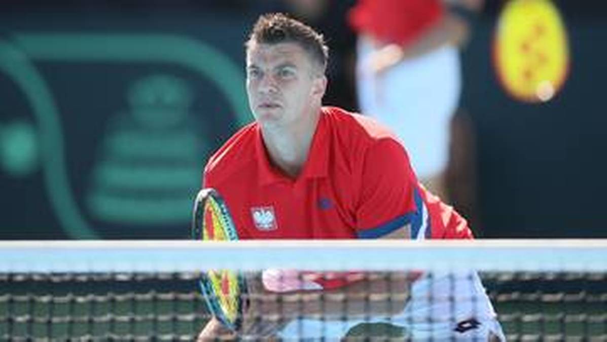 Gracz tenisa z Polski w czerwonej koszulce z orzełkiem, w skupieniu odbija piłkę podczas meczu.