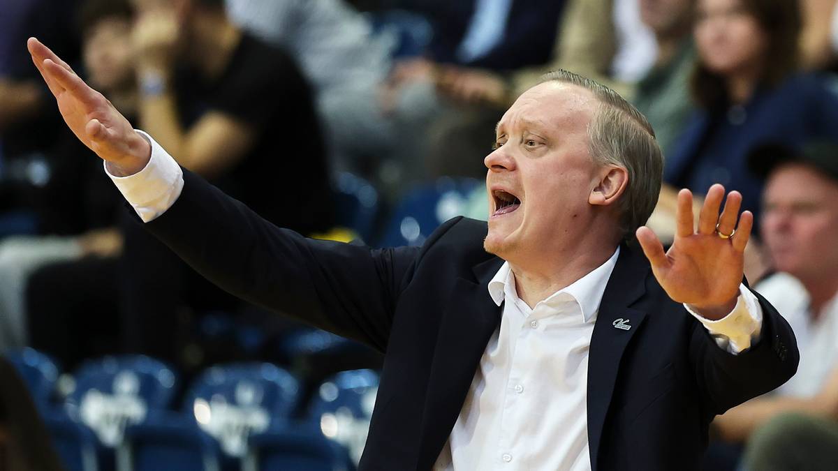 Trener koszykówki w białej koszuli i marynarce, z otwartymi ustami i uniesionymi rękami, wydaje instrukcje.