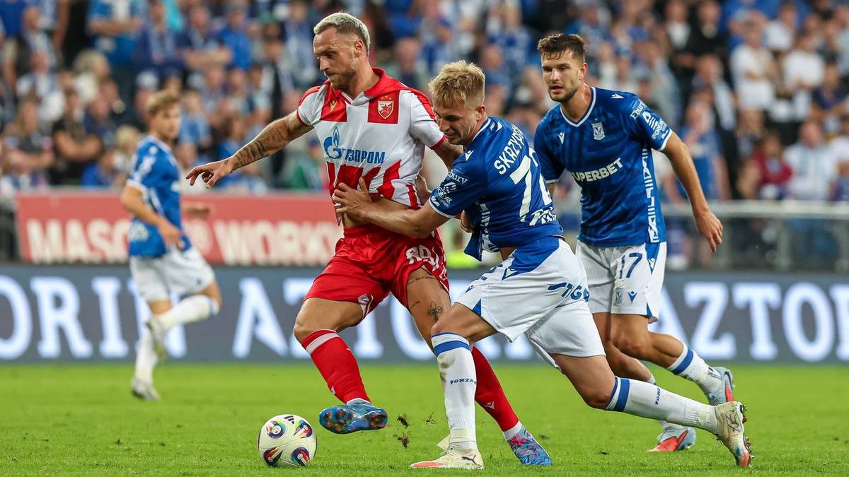 Piłkarz w czerwonej koszulce z białymi elementami walczy o piłkę z dwoma piłkarzami w niebieskich strojach na zielonej murawie stadionu.