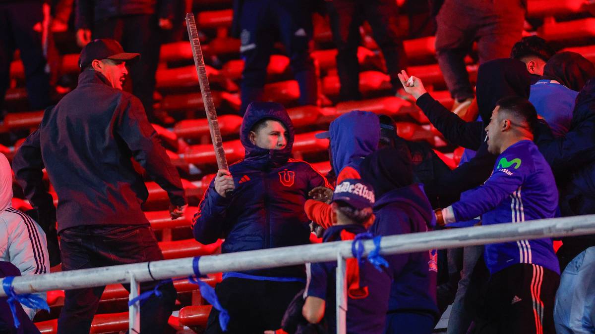 Tłum kibiców na stadionie, jeden z nich trzyma drewniany kij. W tle widać migające światła, sugerujące zamieszki.