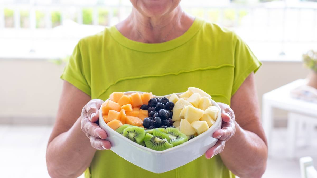 Starsza kobieta trzyma sercowate naczynie z owocami: melon, bor&oacute;wki, kiwi i ananasa.