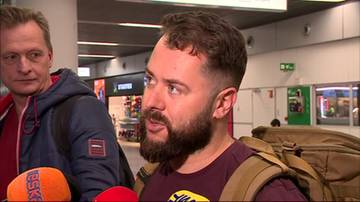 Mężczyzna z brodą i plecakiem na ramieniu, w tle inny mężczyzna i fragment lotniska.