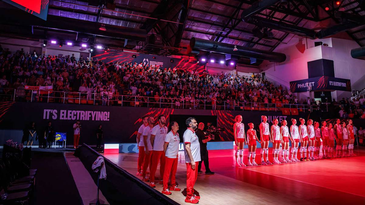 Drużyna siatkarek w białych strojach stoi na boisku przed trybunami wypełnionymi widzami podczas meczu.