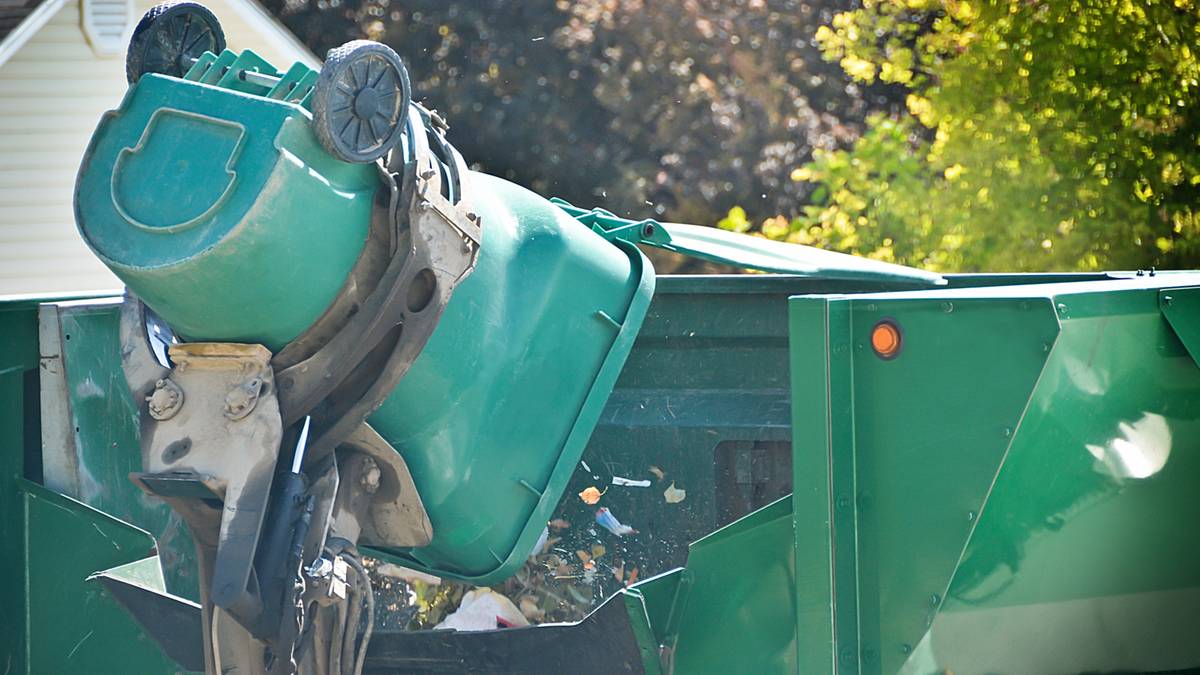 Ramię śmieciarki podnosi zielony kontener na śmieci, wysypując jego zawartość do wnętrza pojazdu.