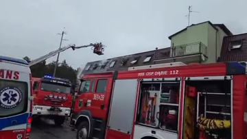 W&oacute;z strażacki z podnośnikiem przy dachu płonącego budynku, widoczny dym i inne wozy strażackie.