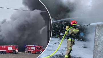 Strażak w pełnym rynsztunku gaśniczym z wężem strażackim na tle płonącej hali przemysłowej i gęstego dymu.