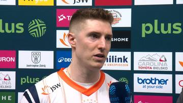 Młody mężczyzna w stroju sportowym, z krótkimi blond włosami, patrzy w prawo z otwartymi ustami. W tle znajduje się ściana z logotypami sponsorów.