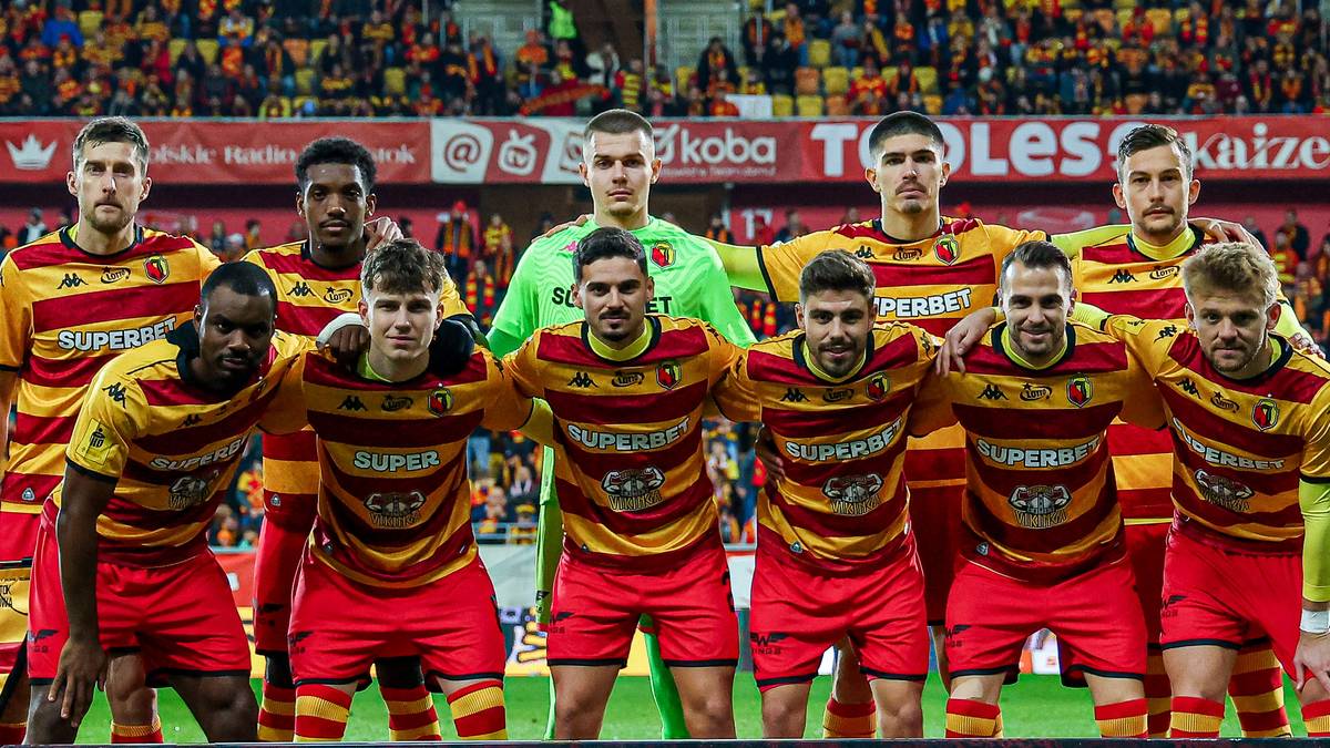 Drużyna piłkarska w czerwonych strojach z żółtymi elementami pozuje do zdjęcia na murawie stadionu.