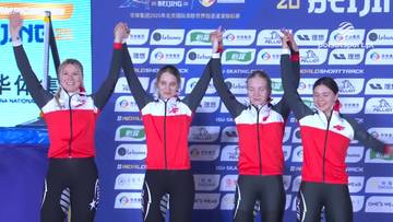 Cztery kobiety w strojach sportowych z czerwonymi i białymi elementami stoją na podium, trzymając się za ręce i świętując sukces.