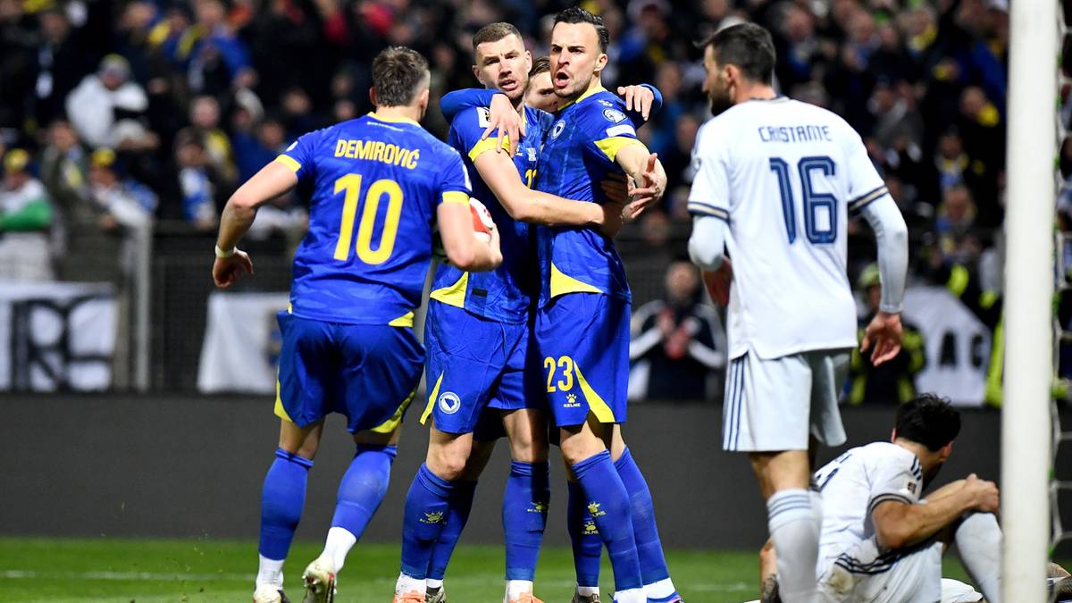 Piłkarze Bośni i Hercegowiny w niebieskich strojach z ż&oacute;łtymi numerami przytulają się po zdobyciu bramki, jeden z nich ma numer 10. Po drugiej stronie boiska stoi piłkarz w białej koszulce z numerem 16, a obok niego klęczy zawodnik w białym stroju bramkarskim.