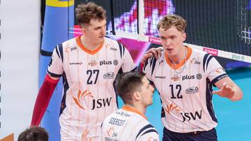 Siatkarze Jastrzębskiego Węgla w białych koszulkach z pomarańczowymi elementami rozmawiają na boisku.