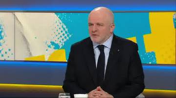 Łysy mężczyzna w czarnym garniturze i białej koszuli z krawatem siedzi i m&oacute;wi, na tle kolorowej grafiki telewizyjnej.