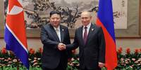 Kim Dzong Un i Władimir Putin ściskają sobie dłonie na tle flag Korei P&oacute;łnocnej i Rosji.