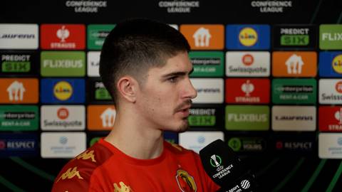 Młody mężczyzna w czerwonej koszulce sportowej z ż&oacute;łtymi detalami, profilowe ujęcie, odpowiada na pytania dziennikarza przed ścianą z logotypami sponsor&oacute;w.