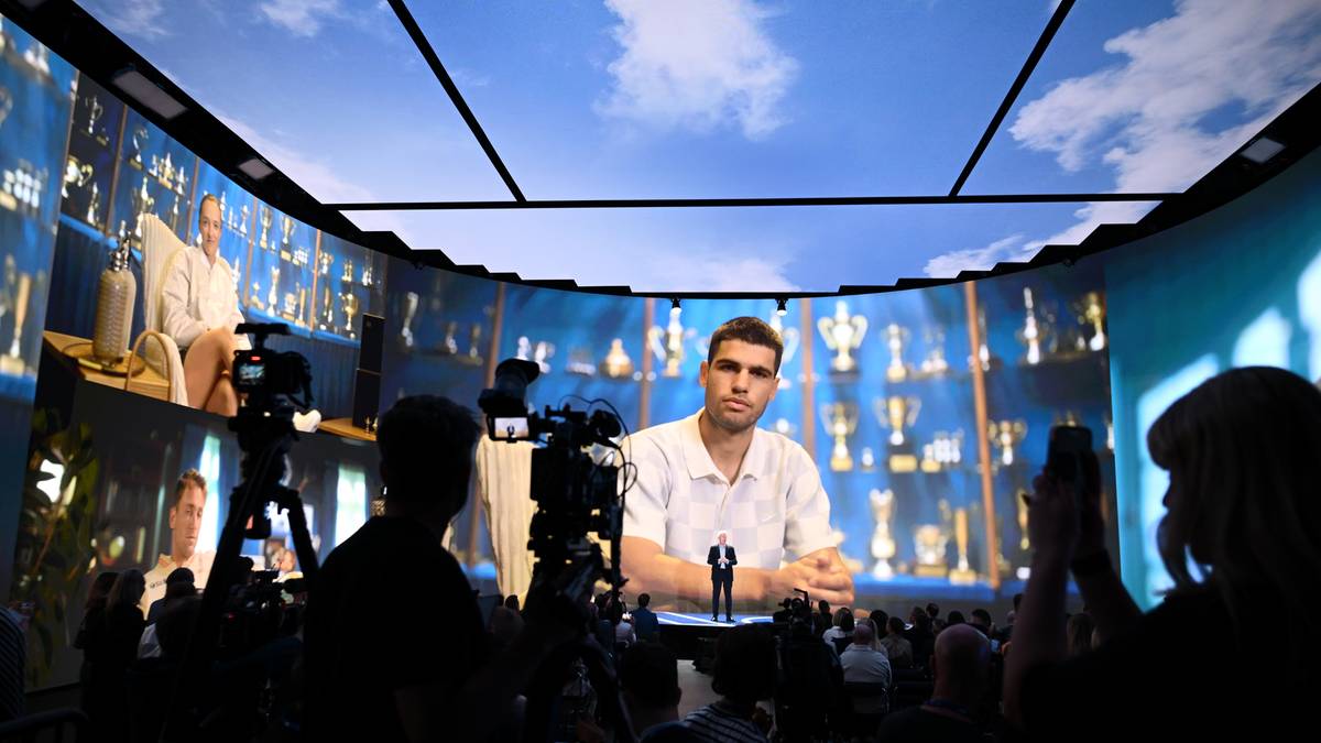 Młody mężczyzna w białej koszulce stoi na podwyższeniu na scenie, przed nim mikroskopijna figura ludzika, a za nim wyświetlana jest gigantyczna projekcja z trofeami i wizerunkiem siedzącego mężczyzny.