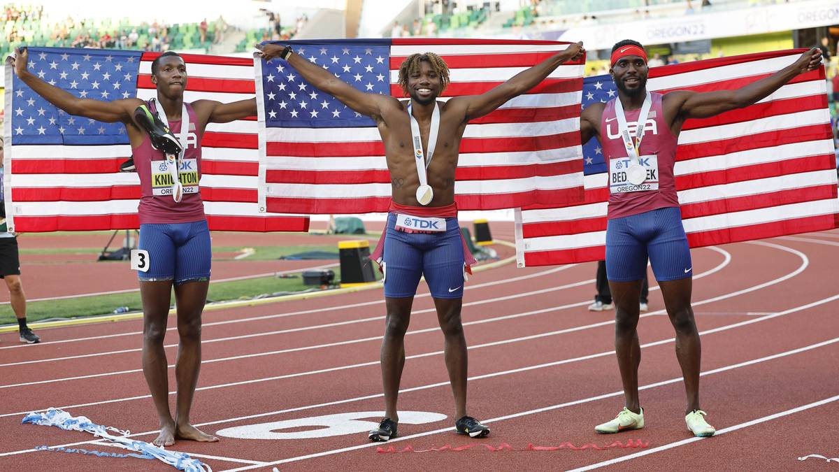 Trzech amerykańskich sprinter&oacute;w na podium z medalami na szyjach, stojących przed flagami USA.