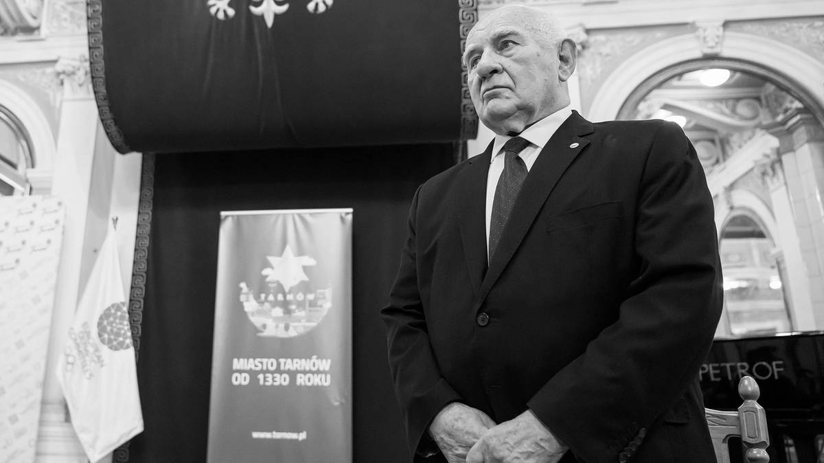 Starszy mężczyzna w garniturze stoi w uroczystym pomieszczeniu.