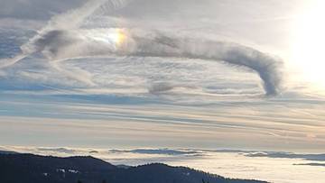 Smuga kondensacyjna samolotu tworząca łuk nad morzem chmur, z widocznym tęczowym efektem świetlnym.