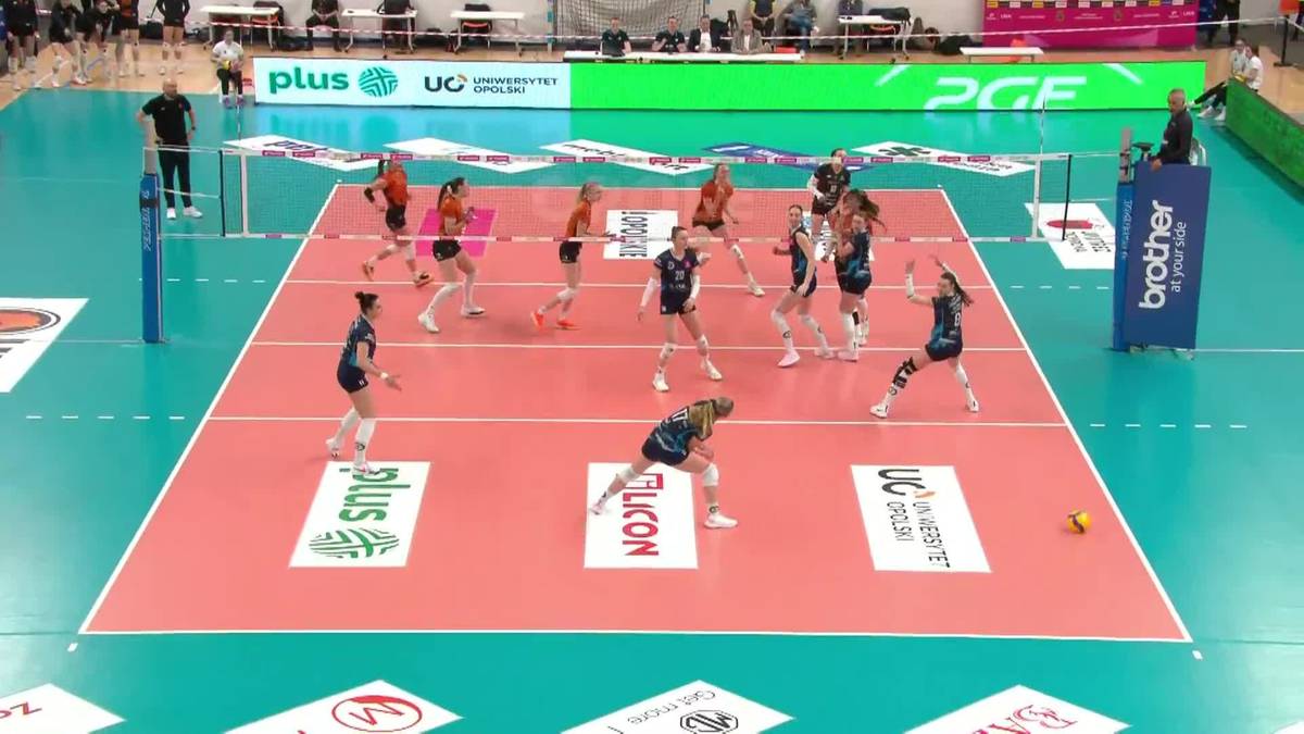 Dwie drużyny siatkarek rywalizują na boisku sportowym, jedna drużyna w niebieskich strojach, druga w białych.