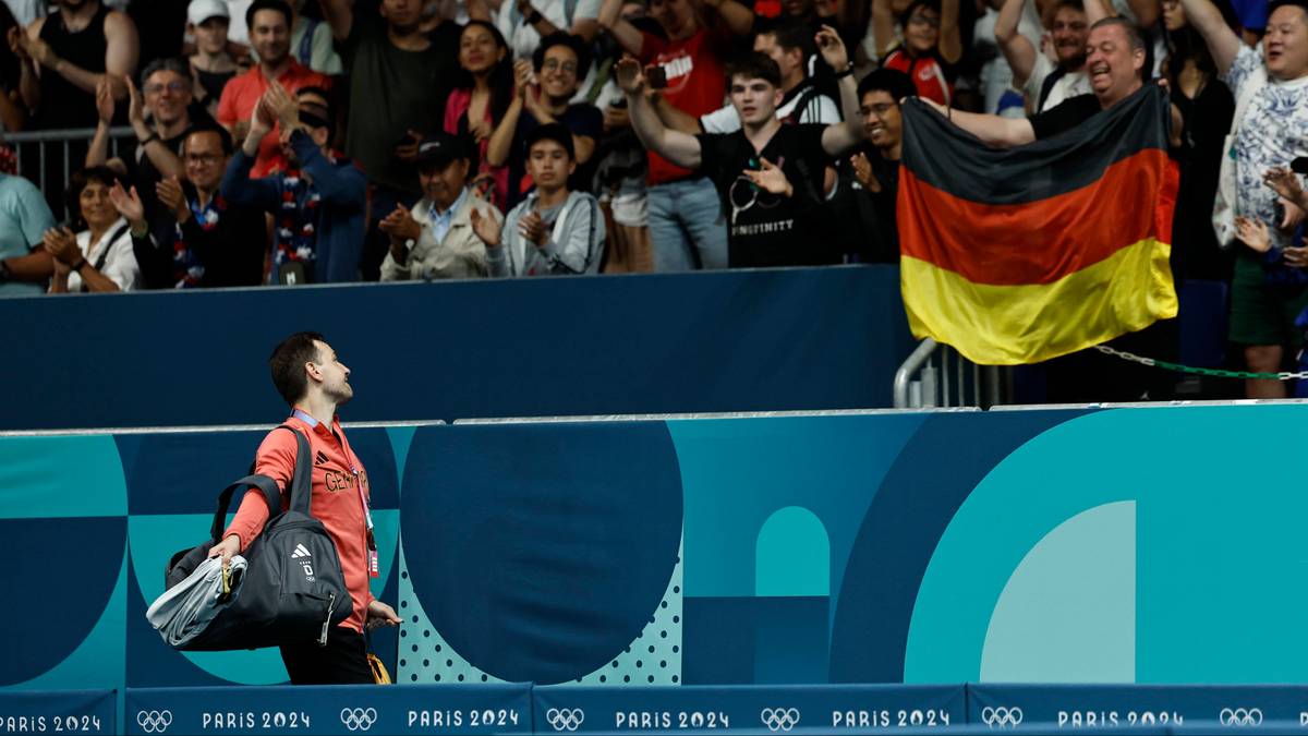 Piłkarz w pomarańczowej koszulce z torbą na ramieniu odwraca się w stronę trybun, na których widzowie trzymają niemiecką flagę.