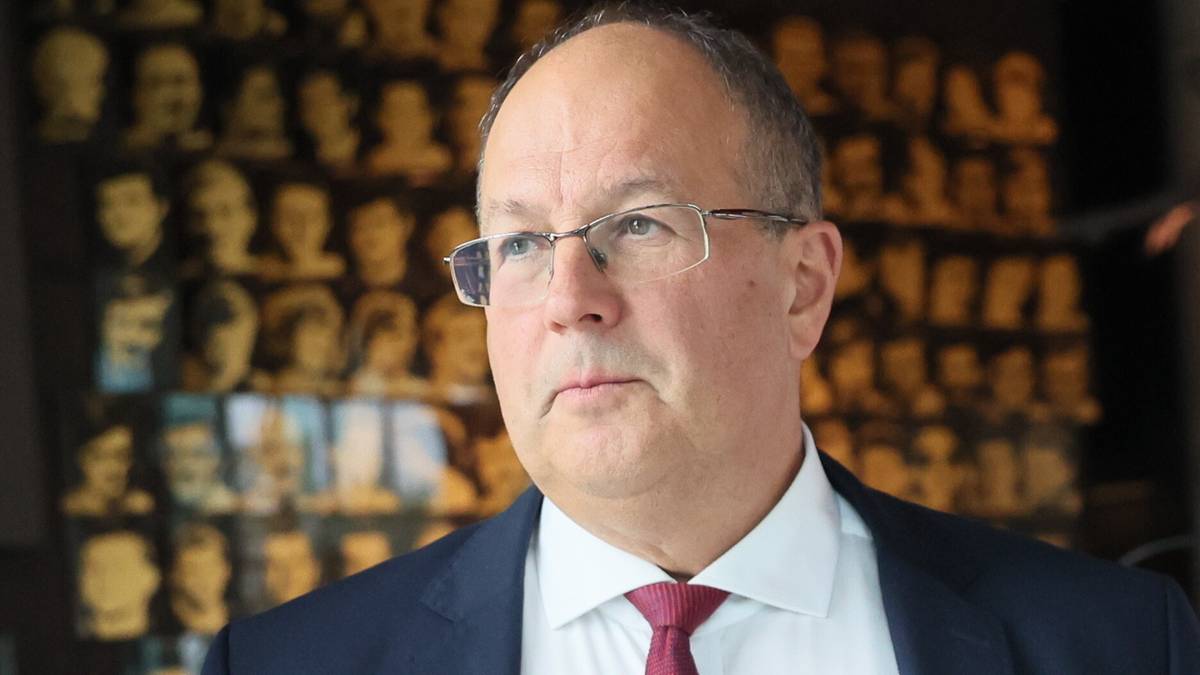 Mężczyzna w okularach i ciemnym garniturze z czerwonym krawatem, ubrany jest w białą koszulę. Jest on na pierwszym planie, a za nim w centrum uwagi znajduje się ściana ozdobiona licznymi popiersiami osób.