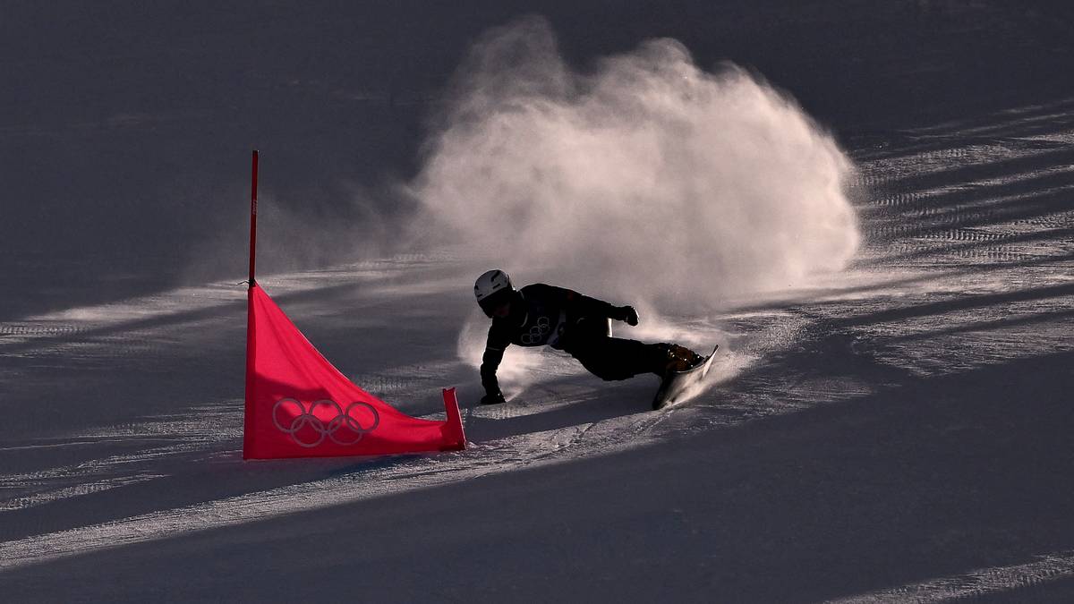 Snowboardzista w akcji na stoku, otoczony mgiełką śniegu, między czerwonymi tyczkami.