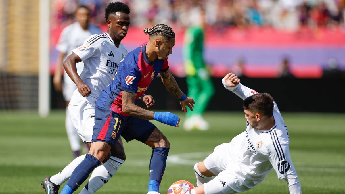 Dwóch piłkarzy w białych strojach Realu Madryt próbuje odebrać piłkę piłkarzowi w niebiesko-czerwonym stroju FC Barcelony. Jeden z zawodników Realu Madryt jest na ziemi, próbując zatrzymać akcję.