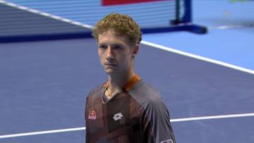 Młody mężczyzna, tenisista, stoi na niebieskim korcie tenisowym, ubrany w szary T-shirt z pomarańczowymi elementami i logo Lotty.