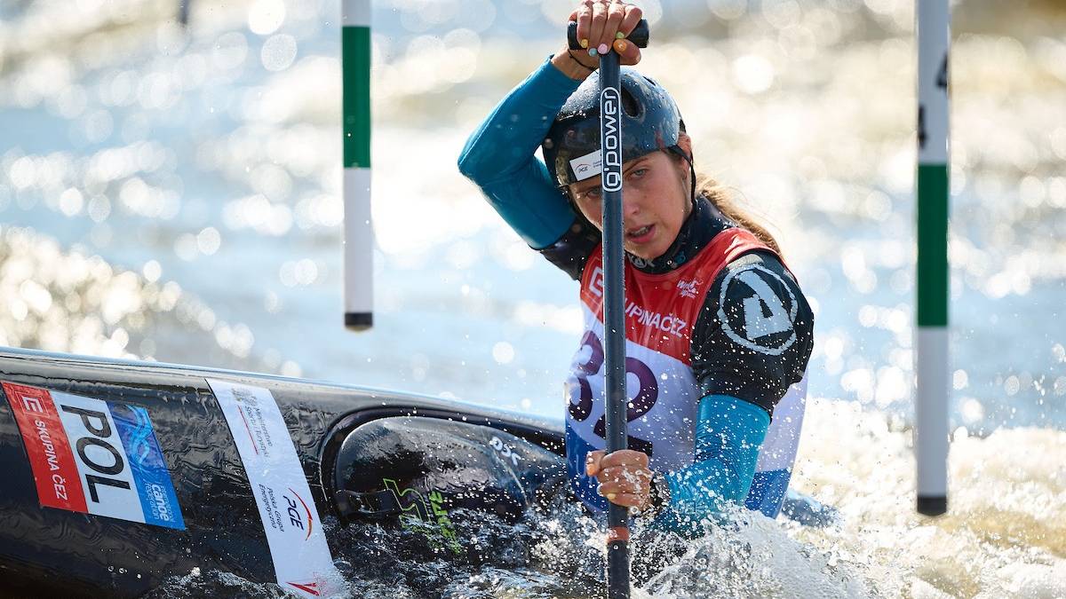 Kobieta w kasku i kamizelce ratunkowej płynie kajakiem, wiosłując w trakcie zawodów slalomu kanadyjkowego.