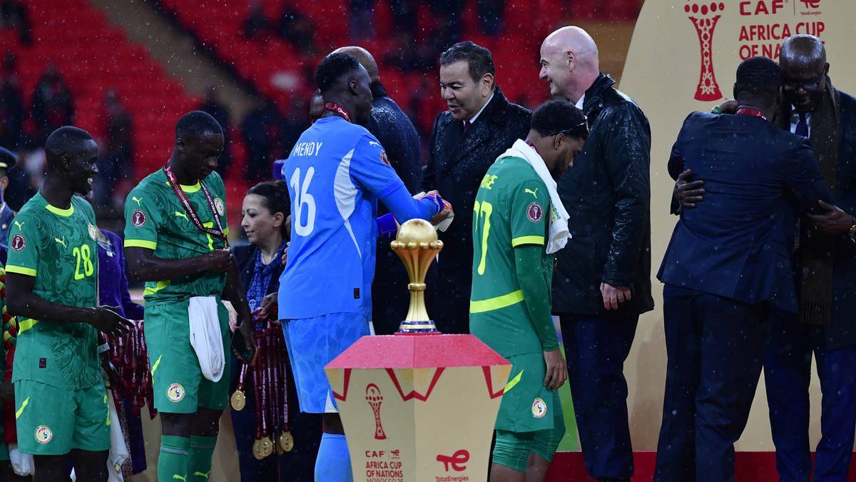 Zawodnicy Senegalu otaczają trofeum, niekt&oacute;rzy wyglądają na przygnębionych, w tle stoją mężczyźni w garniturach.
