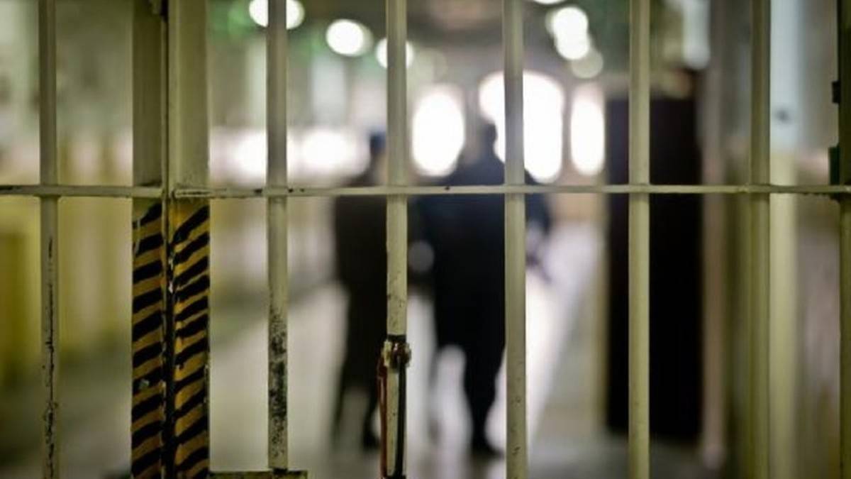 Widok przez więzienne kraty na korytarz z sylwetkami dw&oacute;ch os&oacute;b w tle.