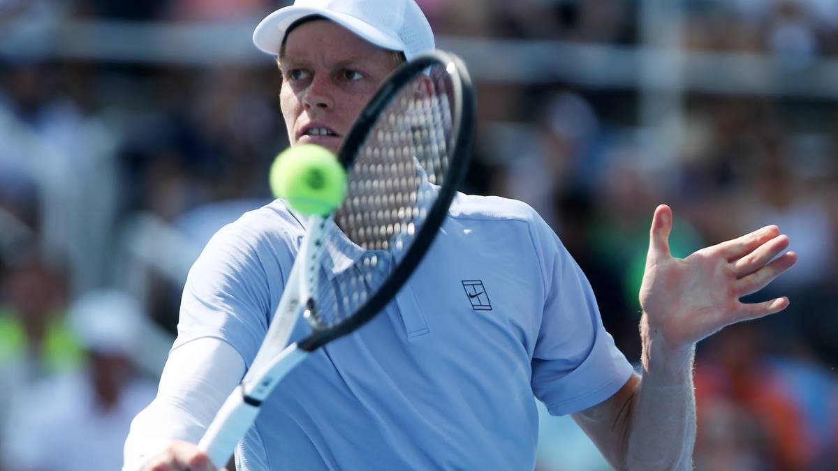 Gracz tenisa w niebieskiej koszulce i białej czapce uderza rakietą piłkę tenisową.