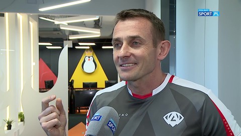 Mężczyzna w szarej bluzie sportowej z białymi wstawkami, udziela wywiadu. W tle widoczne jest nowoczesne pomieszczenie z logo pingwina.