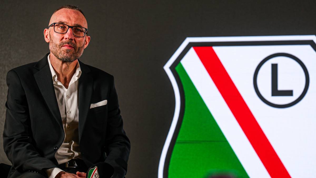 Mężczyzna w okularach siedzi obok herbu Legii Warszawa, trzymając mikrofon.