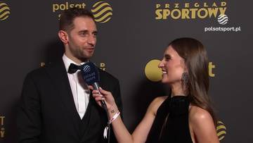 Grzegorz Łomacz, polski siatkarz, w czarnym garniturze z muszką, udziela wywiadu kobiecie z mikrofonem na tle czarnej ściany z logo "Przegląd Sportowy".