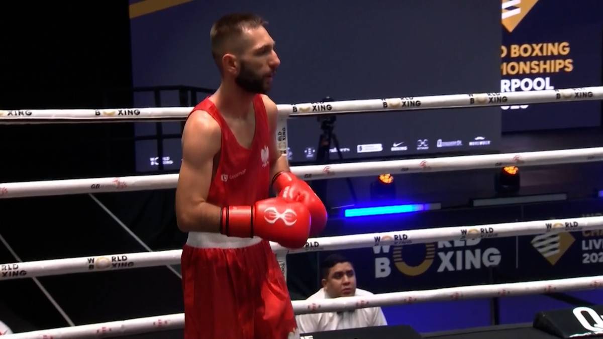 Polski bokser w czerwonym stroju stoi w narożniku ringu, podczas gdy jego rywal znajduje się w tle.