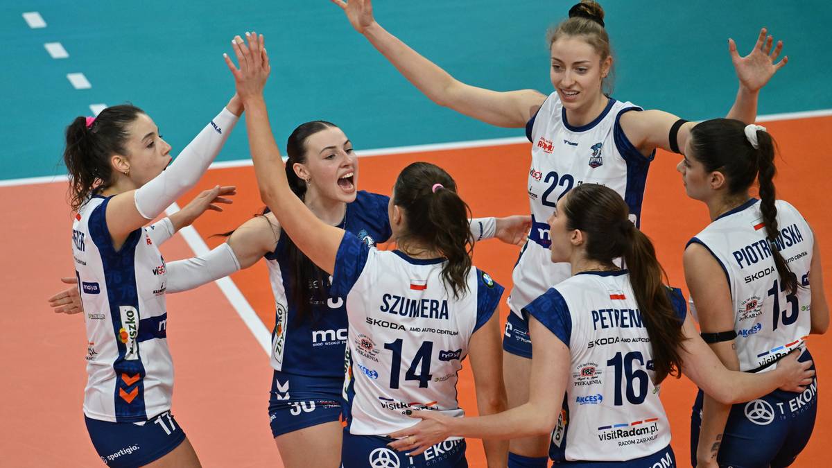Siedem siatkarek w białych strojach z niebieskimi elementami świętuje zwycięstwo na boisku siatkarskim.