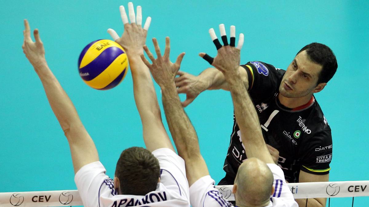 Siatkarze blokuja pileczke w powietrzu. Jeden z nich unosi reke z charakterystycznym kolorem rekawiczki