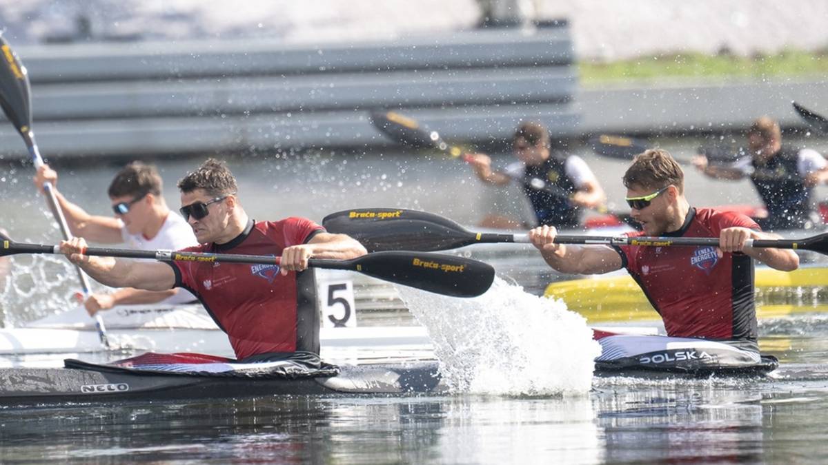 Dwóch kajakarzy w czerwonych koszulkach z numerem 5 ściga się na wodzie, jeden z nich rozpryskuje wodę.